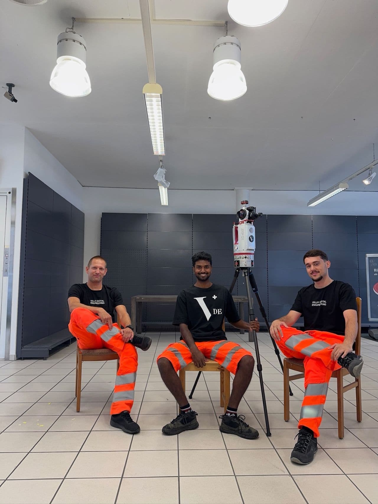 Drei Männer in orangefarbenen Arbeitshosen sitzen auf Stühlen in einem Büro mit grauen Wänden und heller Beleuchtung.