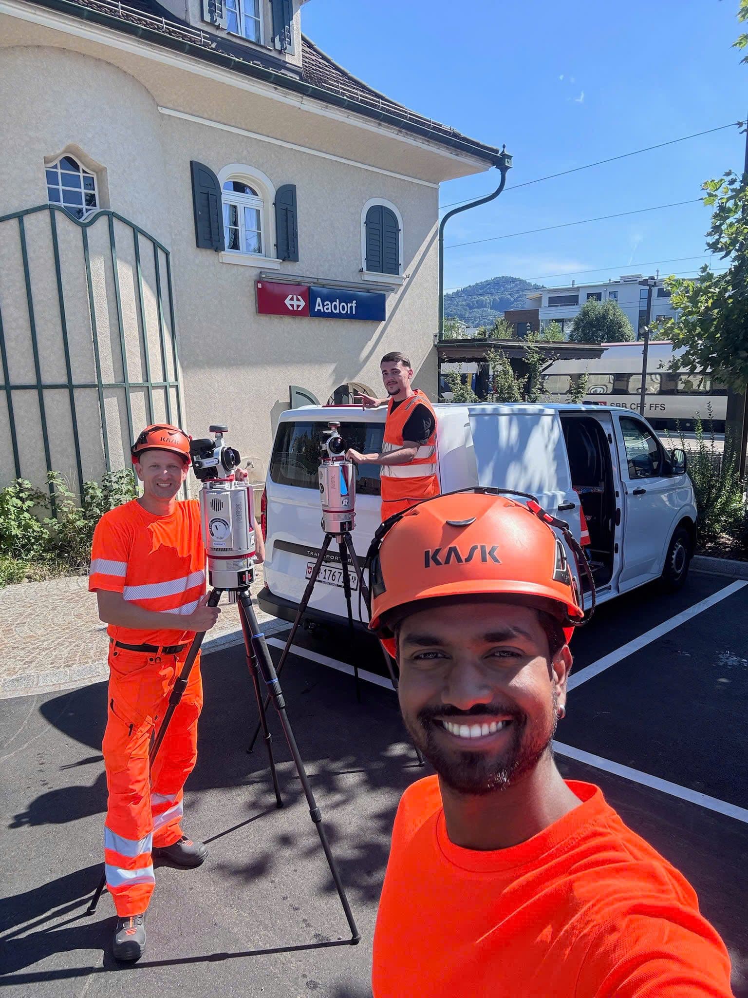 Drei Bauarbeiter in orangefarbener Kleidung arbeiten mit Vermessungsgeräten vor einem weißen Lieferwagen bei sonnigem Wetter. Im Hintergrund ist ein kleines Gebäude mit einem Schild "Aadorf" zu sehen.