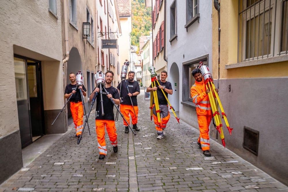 Straßenarbeiter in orangefarbener Schutzkleidung tragen Werkzeuge durch eine enge Gasse.