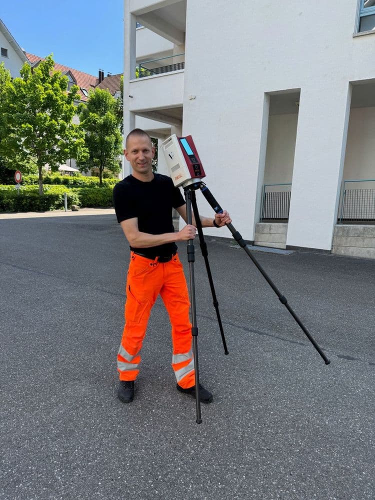 Ein Mann in orangefarbener Arbeitskleidung bedient ein dreibeiniges Vermessungsgerät auf einer Straße vor einem Gebäude.