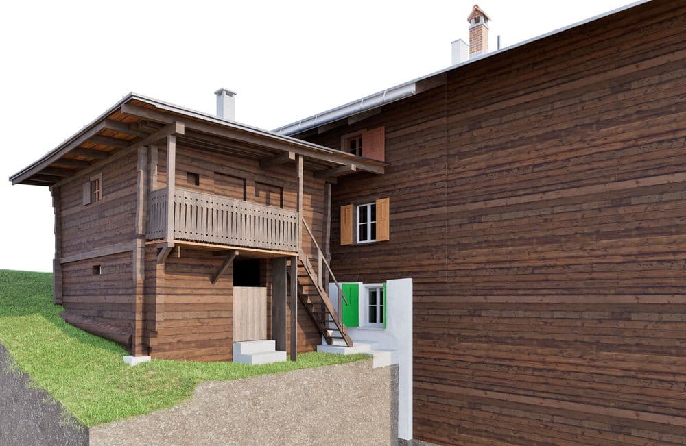 Alpenhaus aus Holz mit Balkon, Treppe und grünem Fensterrahmen auf leicht geneigtem Gelände.