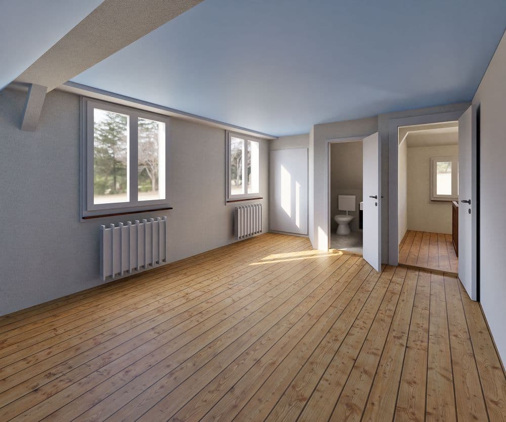 Heller leerer Raum mit Holzfußboden, großen Fenstern, Heizkörpern und zwei offenen Türen, die zu einem Badezimmer führen.