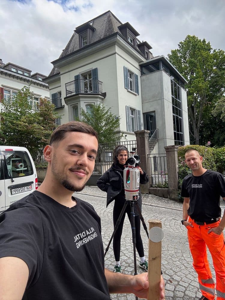 Drei Personen bei Vermessungsarbeiten vor einem großen Gebäude. Eine Person hält eine Messlatte, eine andere bedient ein Vermessungsgerät.