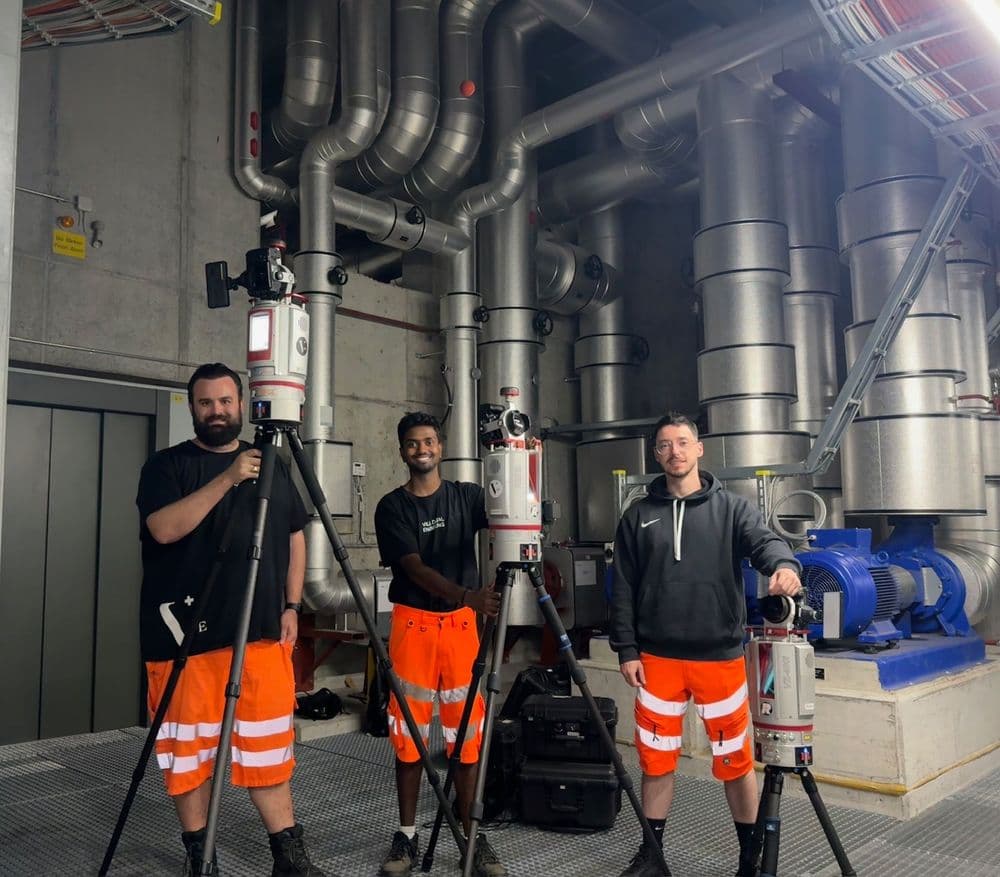 Drei Arbeiter in orangefarbener Schutzkleidung posieren mit Vermessungsgeräten in einer industriellen Anlage. Im Hintergrund sind große Rohre zu sehen.