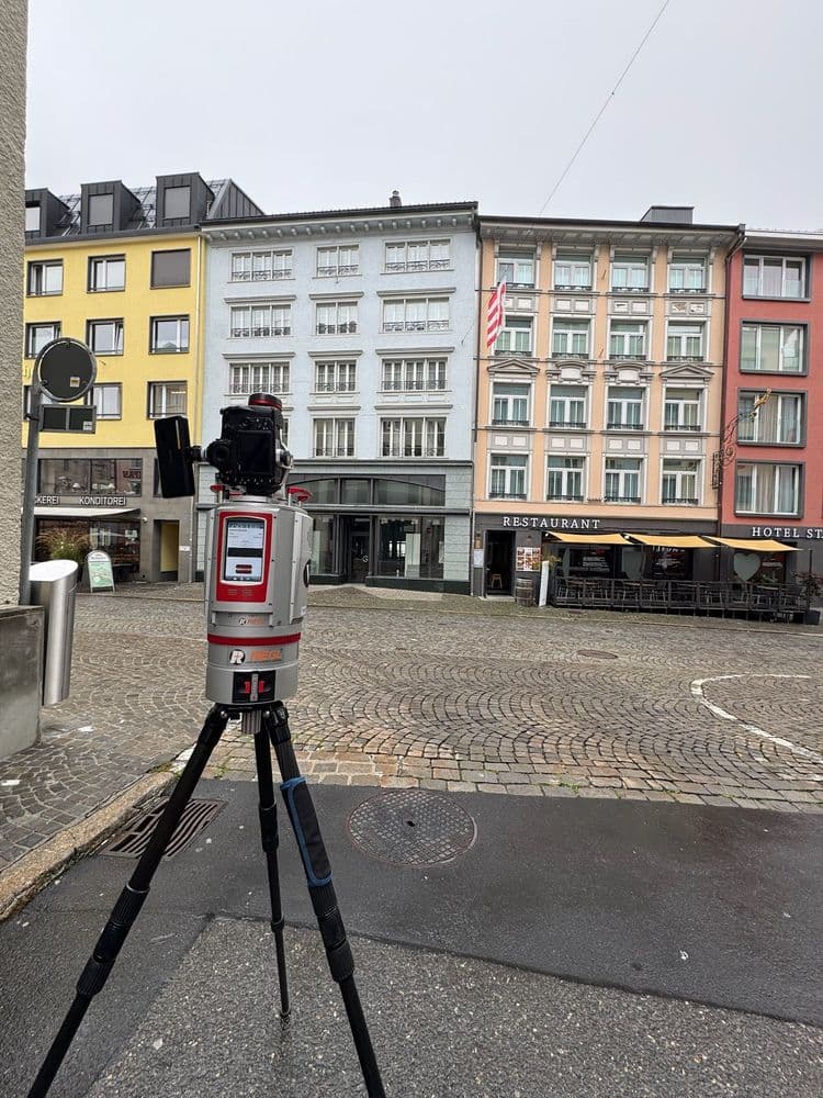 Ein Vermessungsgerät auf einem Stativ steht auf einer gepflasterten Straße vor einer Reihe bunter Gebäude mit Restaurants in einer Stadtumgebung.