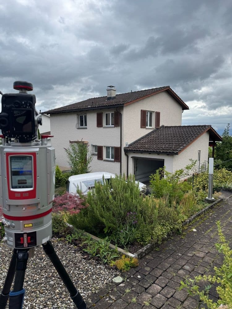 Zweistöckiges Haus mit Garageneinfahrt, umgeben von grünen Pflanzen. Im Vordergrund geodätisches Vermessungsgerät.