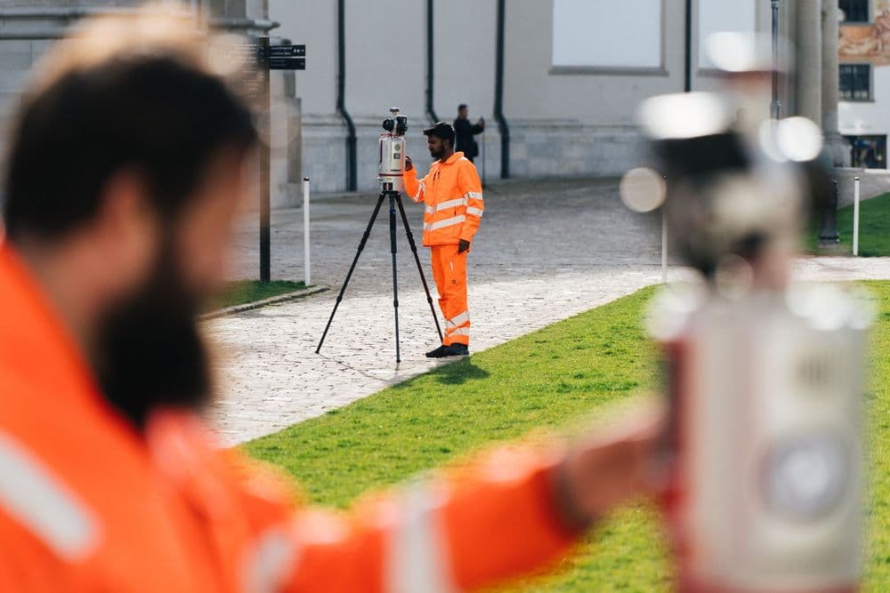 Alternativer Text: "Zwei Landvermesser in orangen Warnwesten arbeiten mit einem Theodolit und einem Stativ im Freien."