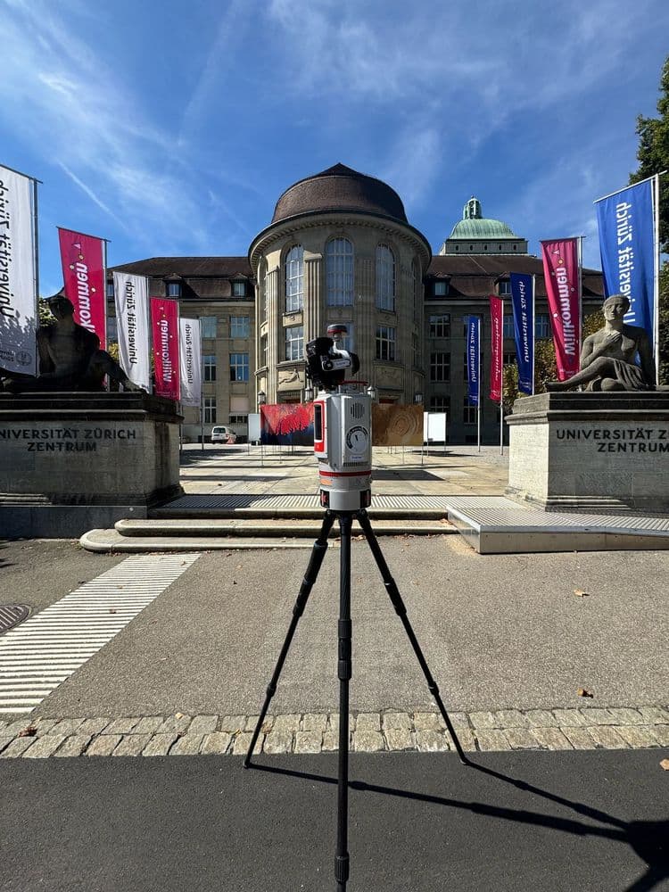 Die Universität Zürich mit zentralem Gebäude, umgeben von Flaggen und Skulpturen, bei sonnigem Wetter. Im Vordergrund ein aufgestelltes weiß-rotes Messgerät auf einem Stativ.