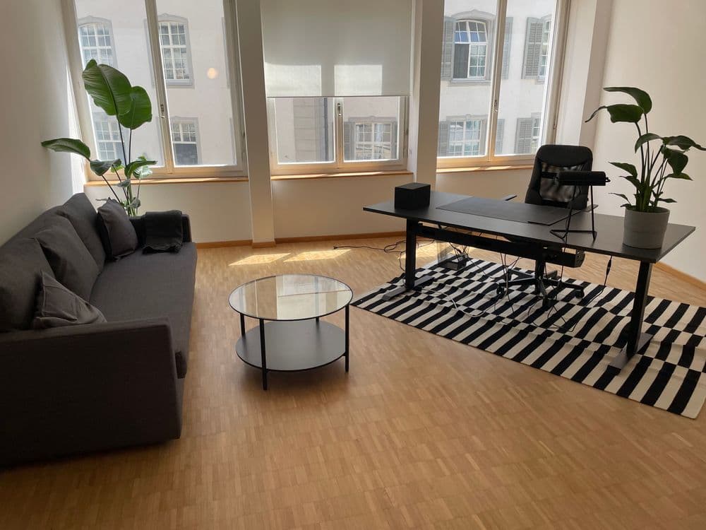 Modernes Büro mit Schreibtisch, Sofa, Couchtisch und Pflanzen, großer Fensterfront, Holzboden und gestreiftem Teppich.