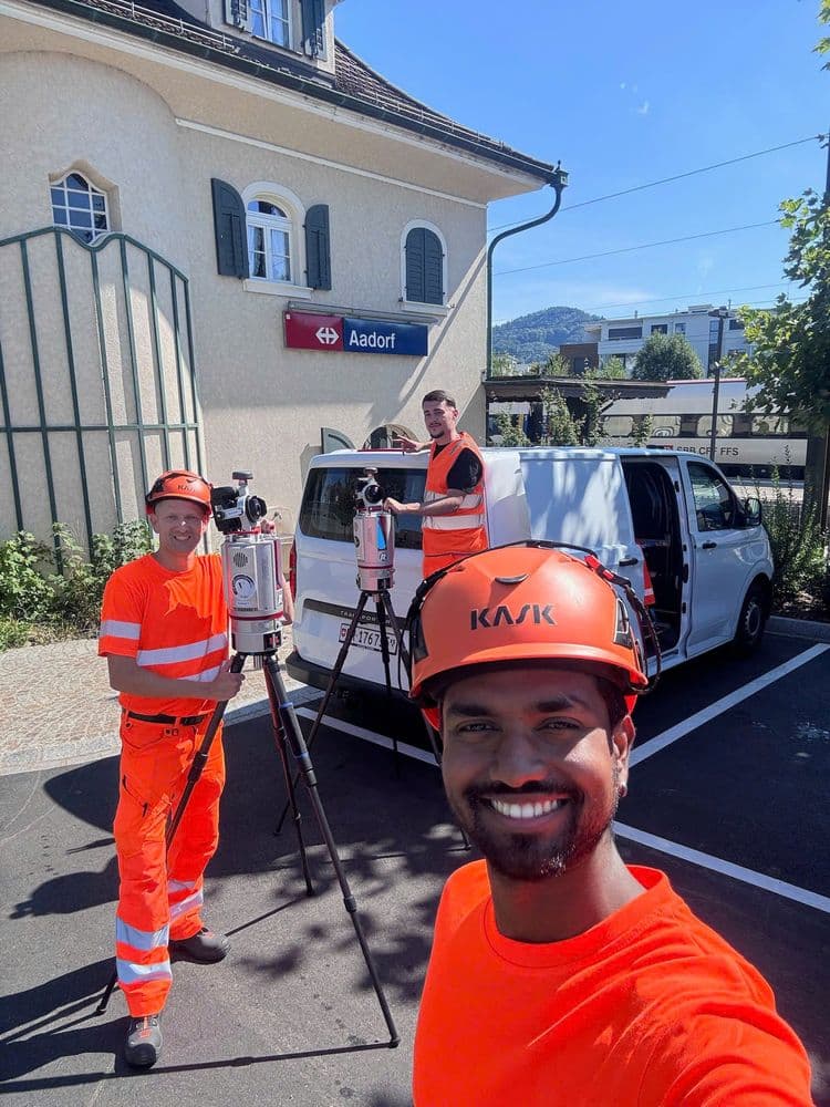 Drei Bauarbeiter in orangefarbener Kleidung arbeiten mit Vermessungsgeräten vor einem weißen Lieferwagen bei sonnigem Wetter. Im Hintergrund ist ein kleines Gebäude mit einem Schild "Aadorf" zu sehen.