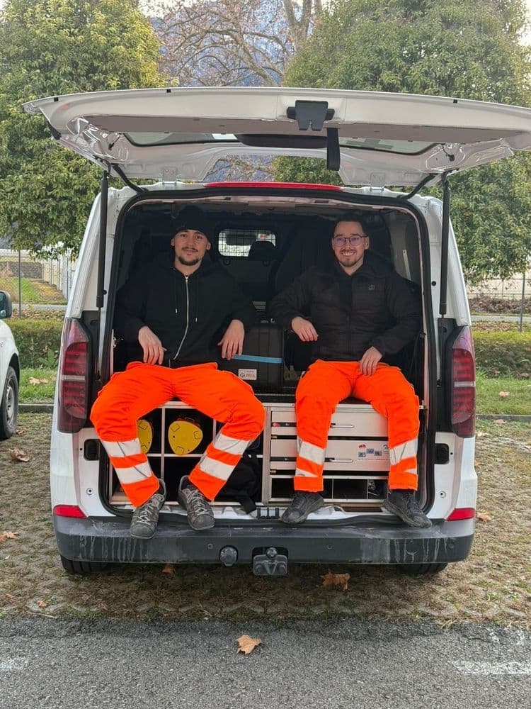 Zwei Männer in oranger Arbeitskleidung sitzen im offenen Kofferraum eines weißen Transporters.