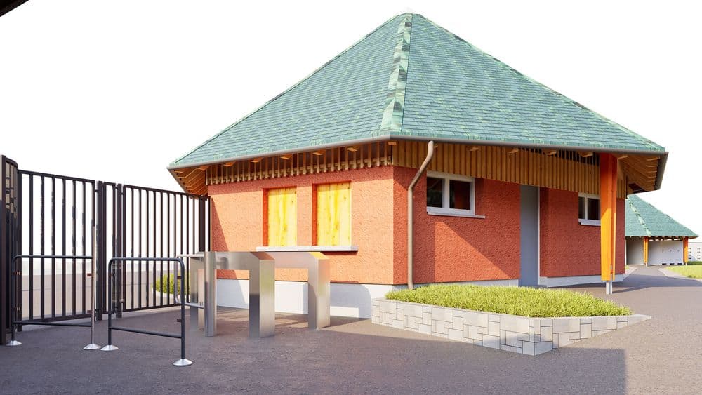 Modernes Backsteingebäude mit grünem Walmdach, kleinen Fenstern und Eingang, davor Metallzaun mit Tor und Pollern; gepflasterter Hof und bepflanztes Beet neben dem Gebäude.