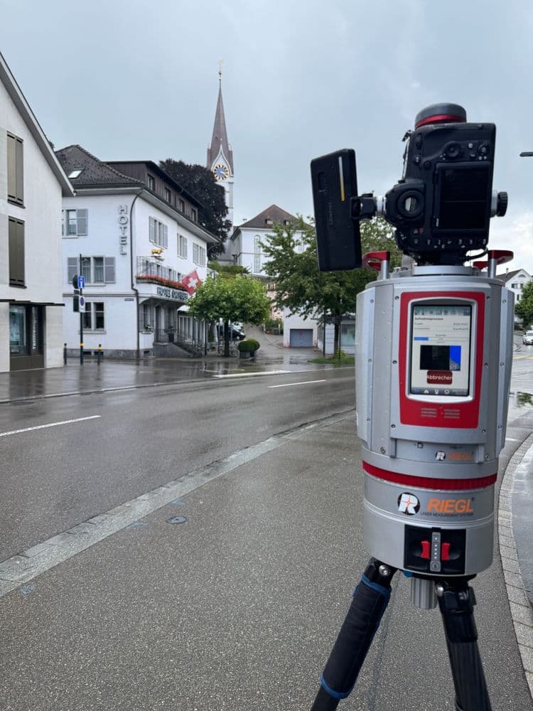 Ein RIEGL 3D-Laserscanner auf einem Stativ erfasst eine Straßenszene mit Gebäuden und einem Kirchturm im Hintergrund.