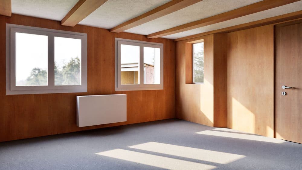 Heller leerer Innenraum mit Holzvertäfelung, sichtbaren Deckenbalken, zwei Fenstern und Wandheizkörper; Sonnenlicht fällt auf grauen Boden, rechts Tür und schmaler Fensterstreifen.