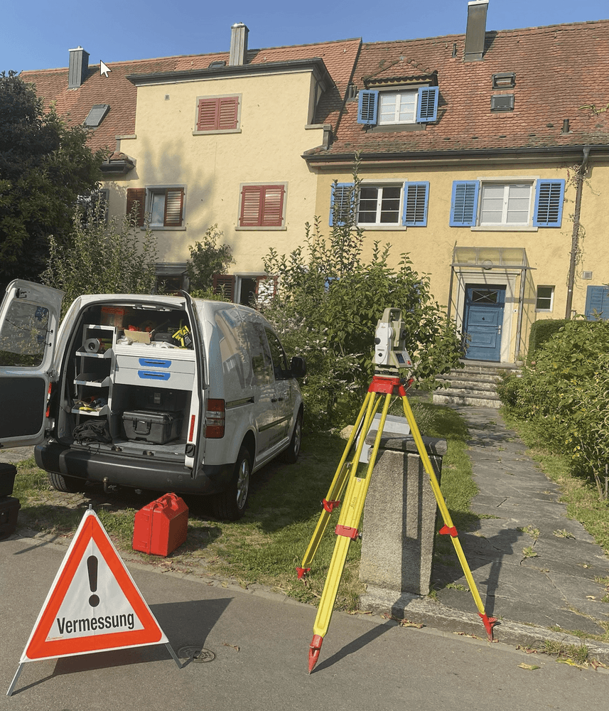 Ein Vermessungstransit steht vor einem Wohngebäude. Ein weißer Lieferwagen mit geöffneter Heckklappe und ein Warnschild "Vermessung" sind sichtbar.