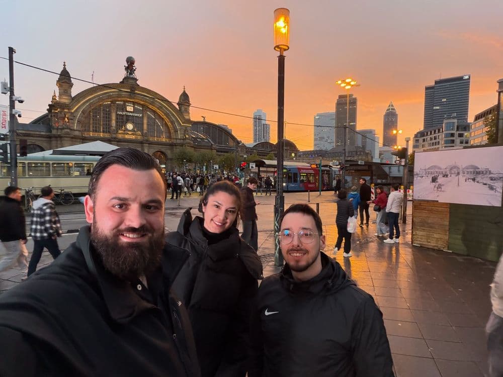 Drei Personen posieren bei Sonnenuntergang vor dem Hauptbahnhof Frankfurt, umgeben von vorbeigehenden Menschen und einer urbanen Kulisse.
